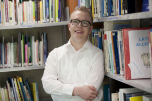 A young man standing in the corner of the library with his arms crossed and smiling at the camera.