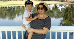 Mother wearing glasses and a hat is holding a young boy who is smiling at the camera