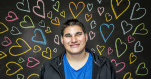 Young man with short dark hair smiles at the camera. There are different coloured hearts drawn on the blackboard in the background.