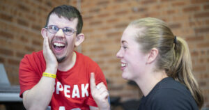 Man with glasses on the left laughs and has hand on the side of his face, with a young woman smiling and pointing her finger up.