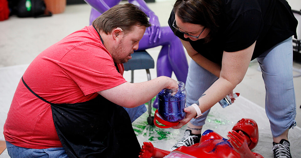 A man with Down syndrome in a red t-shirt adds blue glitter to a red mannequin.