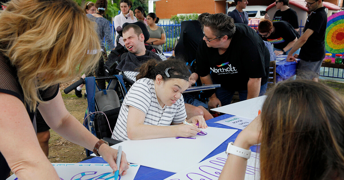 A large group of people with disability sit around tables and write on signs.