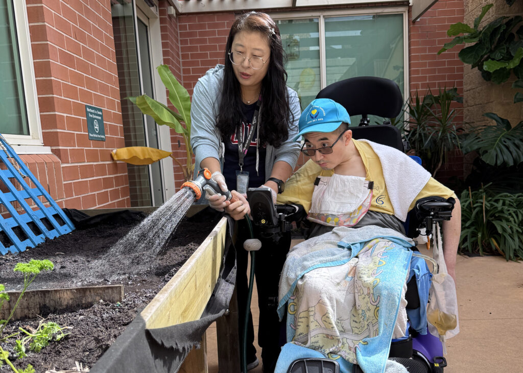 A Northcott support worker is helping a customer water some plants in a raised garden bed