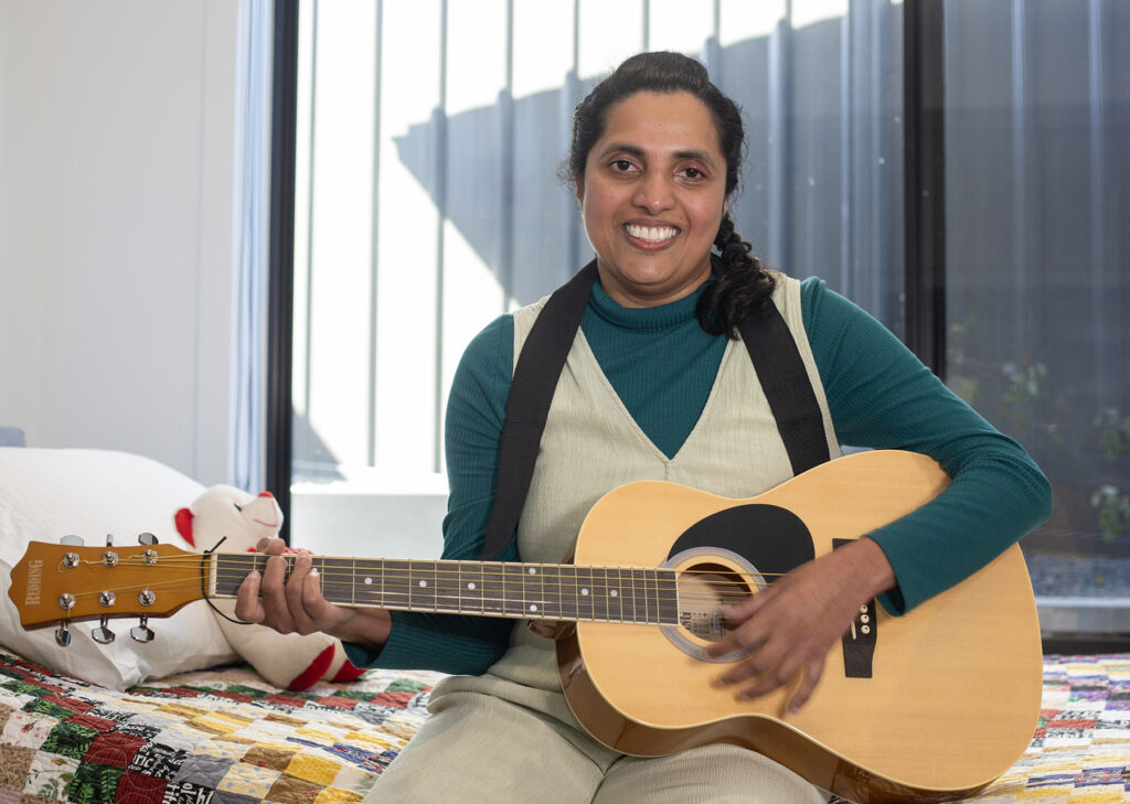 Vindya is playing her guitar in her bedroom - she is smiling happily.