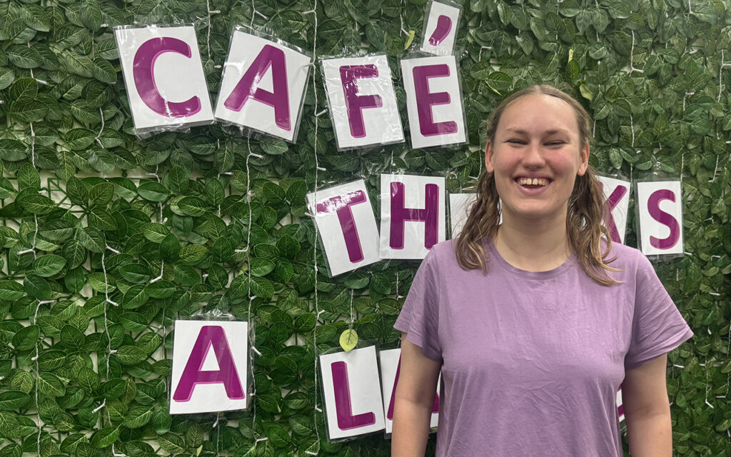 Vanessa is smiling in this photo. She is standing in front of a green wall that has a sign saying