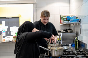 Two people in a commercial kitchen are adding items to a pan