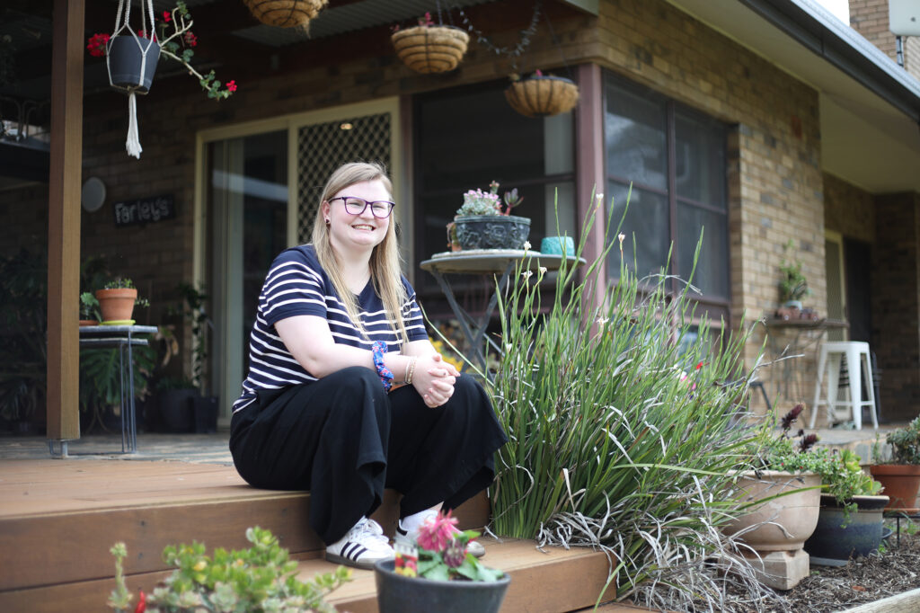 A young woman with long blonde hair wearing glasses is sitting on an outside step smiling at the camera.