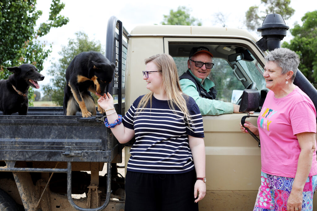 A young woman with long blonde hair is smiling and patting a black dog in the back of a utility vehicle. There is another black dog in the vehicle. Her parents are watching on and smiling.