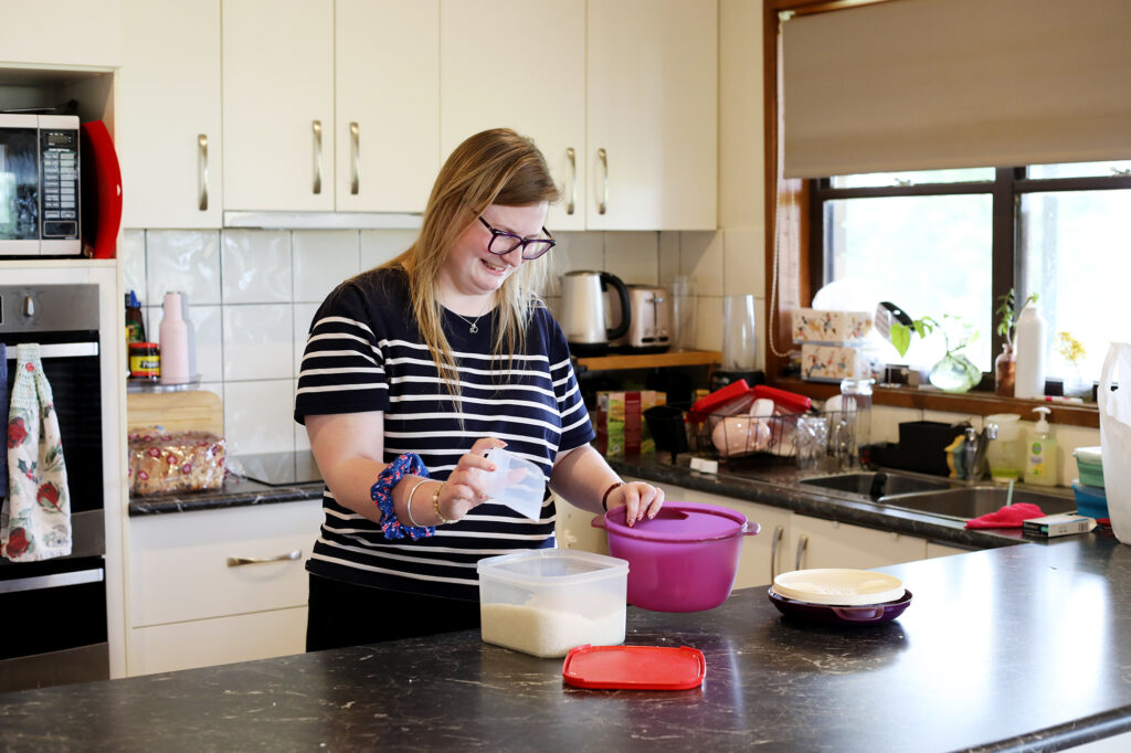 A young woman with long blonde hair and glasses is using a mixing bowl in the kitchen.