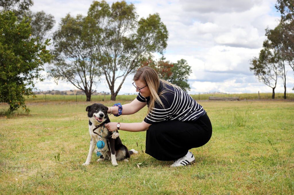 A young woman with long blonde hair and glasses is kneeling down on the grass to pat a dog.