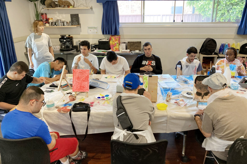 Large group of customers at w long table prepare their work for upcoming art exhibition.