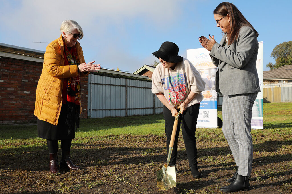 Two people watch another person dig a small hole with a shovel