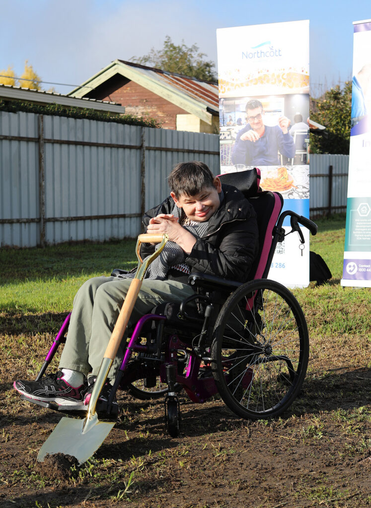 A person in a wheelchair is pictured on a building plot. They have a spade.