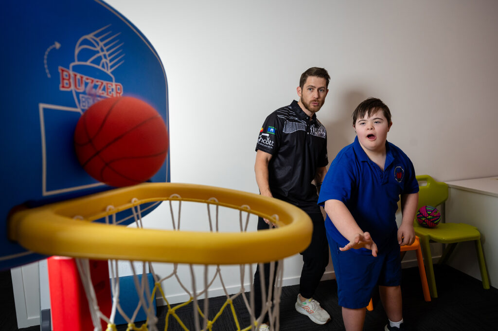 A teenage boy throws a ball into a basket while a Northcott staff member watches.